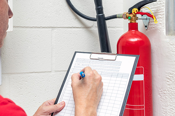 person holding a clipboard while writing next to a fire extinguisher with safety checklist and inspection document for effective fire safety management