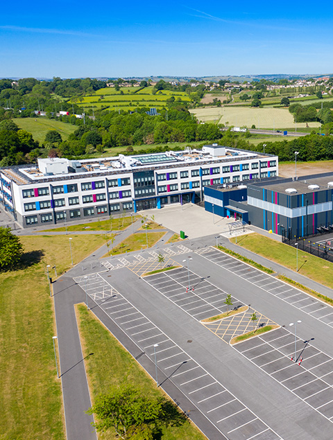 aerial view of a modern school building surrounded by green fields and a large empty parking lot designed for four vehicles