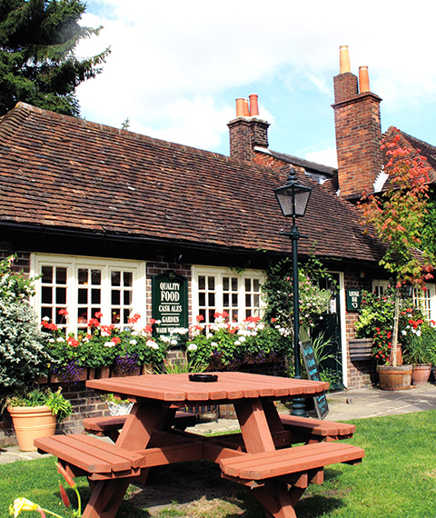 a quaint rustic building with a thatched roof surrounded by vibrant flowers and greenery featuring a wooden picnic table in the foreground perfect for enjoying meals outdoors gardens and relaxation 3 locations to visit
